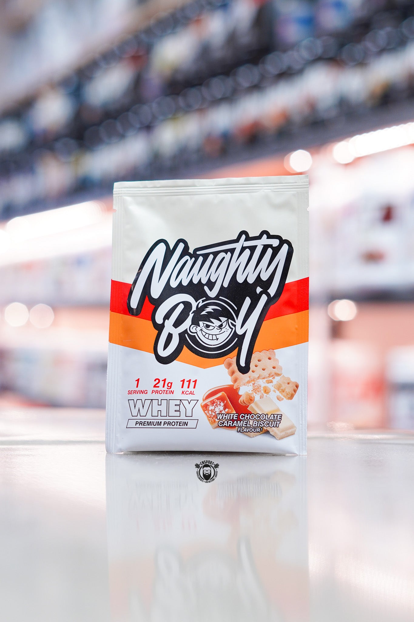 Naughty Boy Whey Protein 30g sample sachet White Chocolate Caramel Biscuit flavour on retail counter with blurred supplement store background