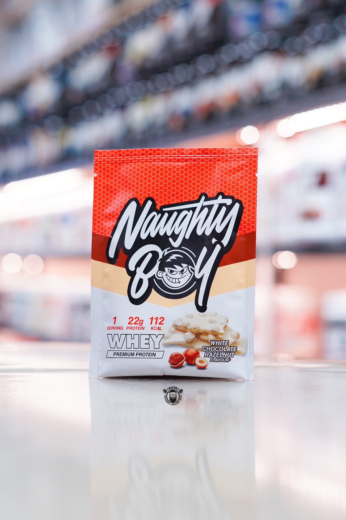 Naughty Boy Whey Protein 30g sample sachet White Chocolate Hazelnut flavour on retail counter with blurred supplement store background