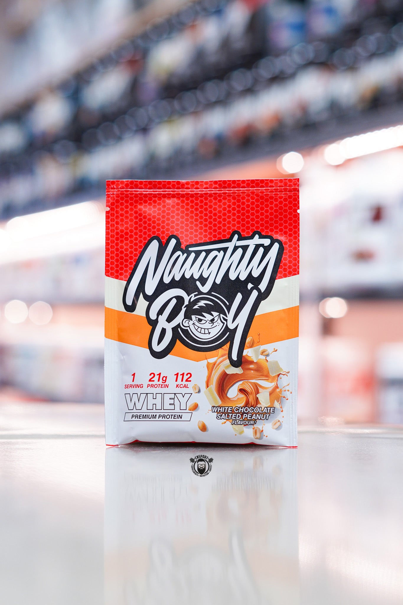 Naughty Boy Whey Protein 30g sample sachet White Chocolate Salted Peanut flavour on retail counter with blurred supplement store background