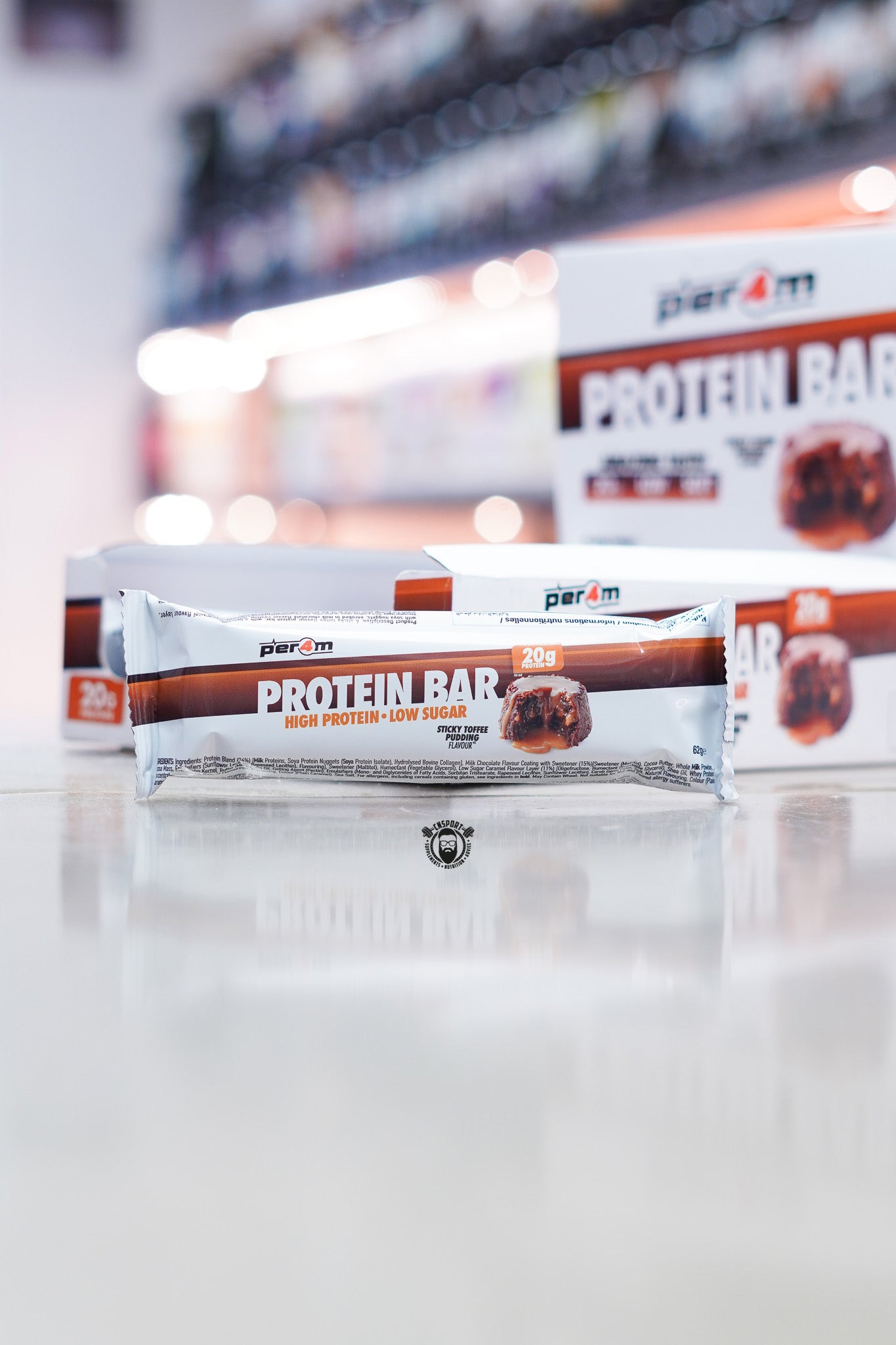 Per4m Protein Bar Sticky Toffee Pudding flavour with 20g protein and low sugar displayed in front of Per4m retail display boxes on supplement store counter with blurred background