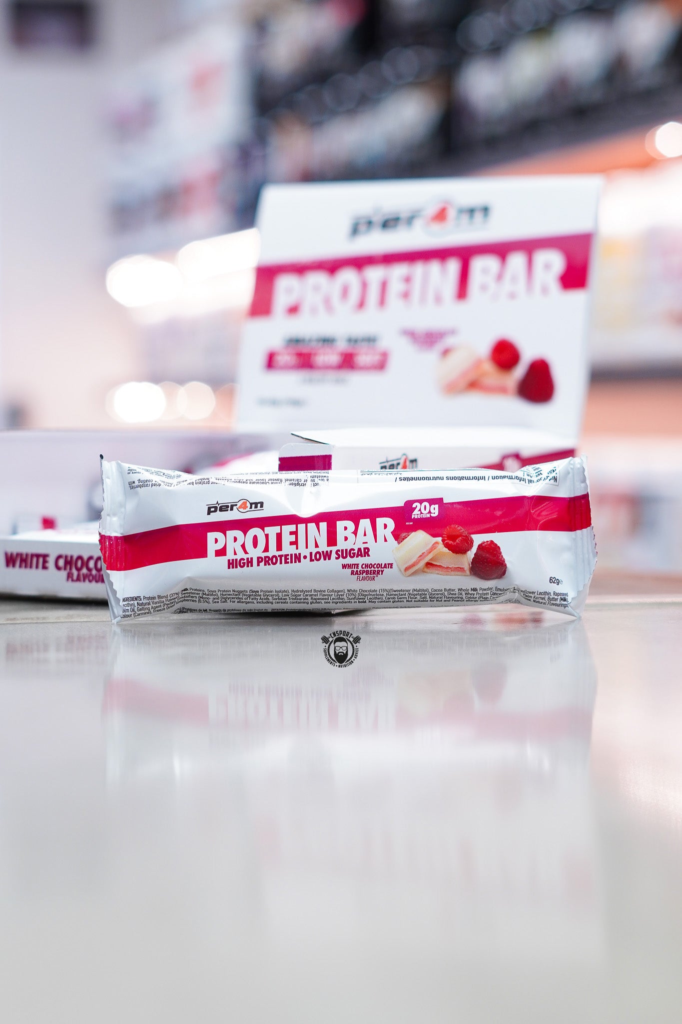 Per4m Protein Bar White Chocolate Raspberry flavour displayed on supplement store counter with product box in the background, highlighting 20g protein, high protein and low sugar formulation, with white chocolate and raspberry flavour imagery.