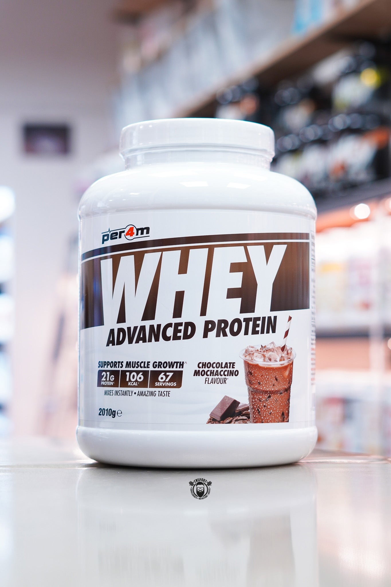 Per4m Whey Advanced Protein Chocolate Mochaccino 2kg protein powder displayed on a countertop in a supplement store, featuring 21g protein per serving, 106 calories, 67 servings, and a chocolate iced coffee flavour graphic on the front label.