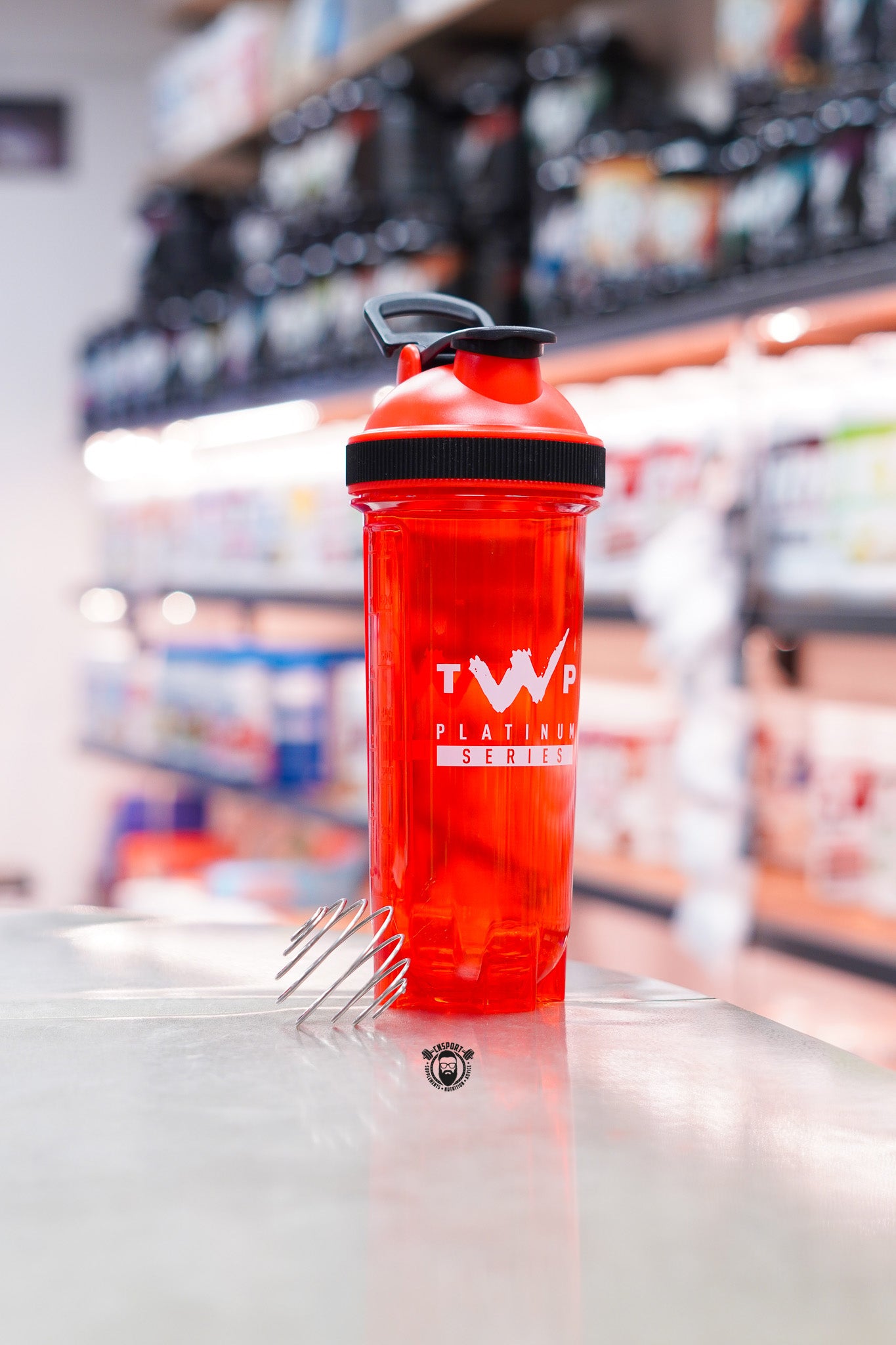 TWP Platinum Series 800ml red blender ball shaker with stainless steel mixing whisk, displayed on a supplement store counter.