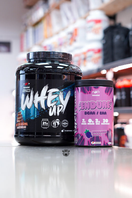 TWP All The Whey Up protein powder, Salted Caramel Shortbread flavour, alongside TWP Endure BCAA/EAA Bubblegum Grape tub, displayed on counter in supplement store