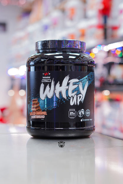 TWP Platinum Series All The Whey Up protein powder in Salted Caramel Shortbread flavour, 900g tub shown on a countertop with blurred supplement shelves behind.