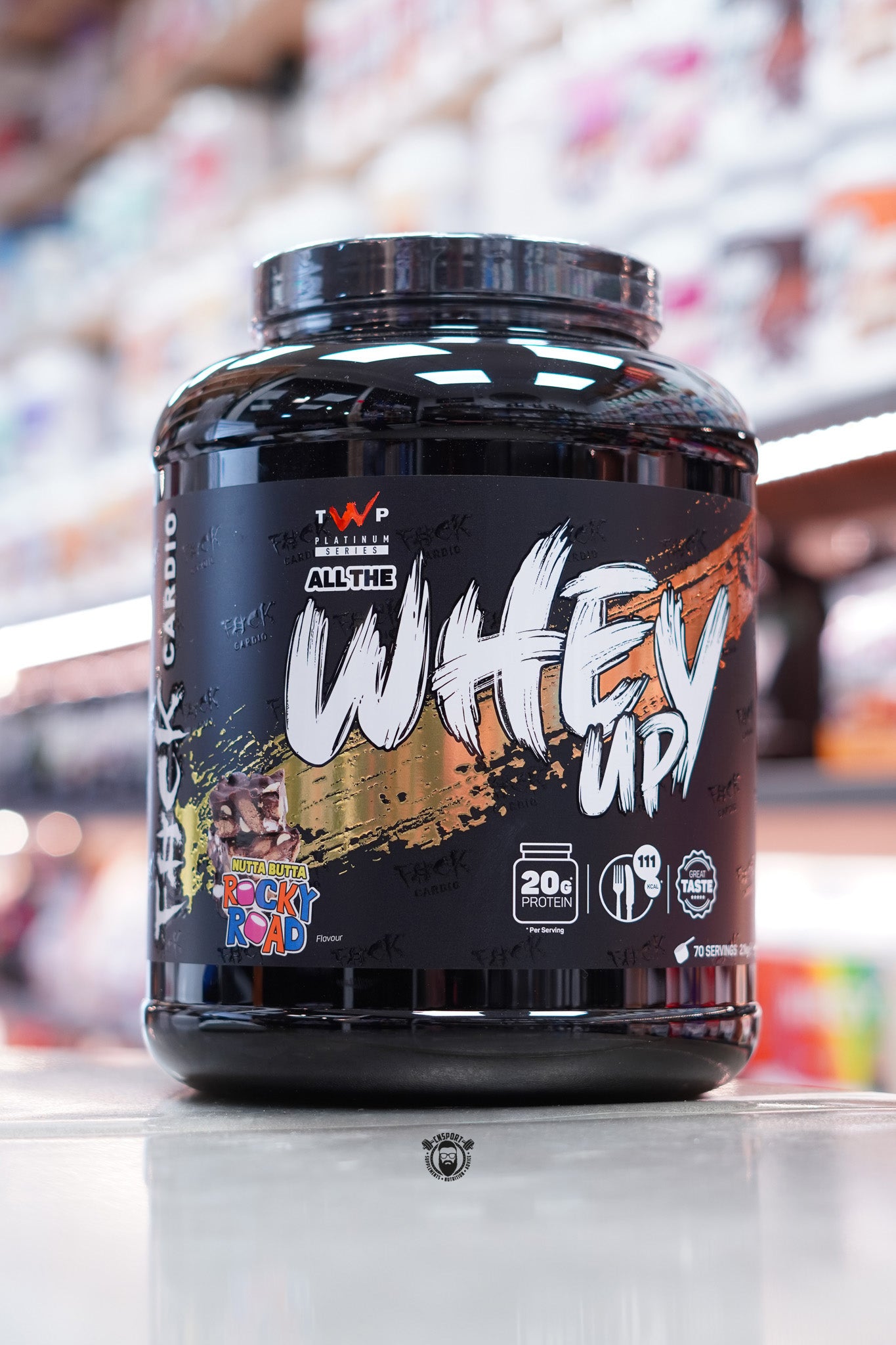 Large black tub of TWP All The Whey Up protein powder in Nutta Butta Rocky Road flavour, with white and gold text on a dark label. Design includes an image of a rocky road dessert and callouts for 20g protein and 111 calories per serving.