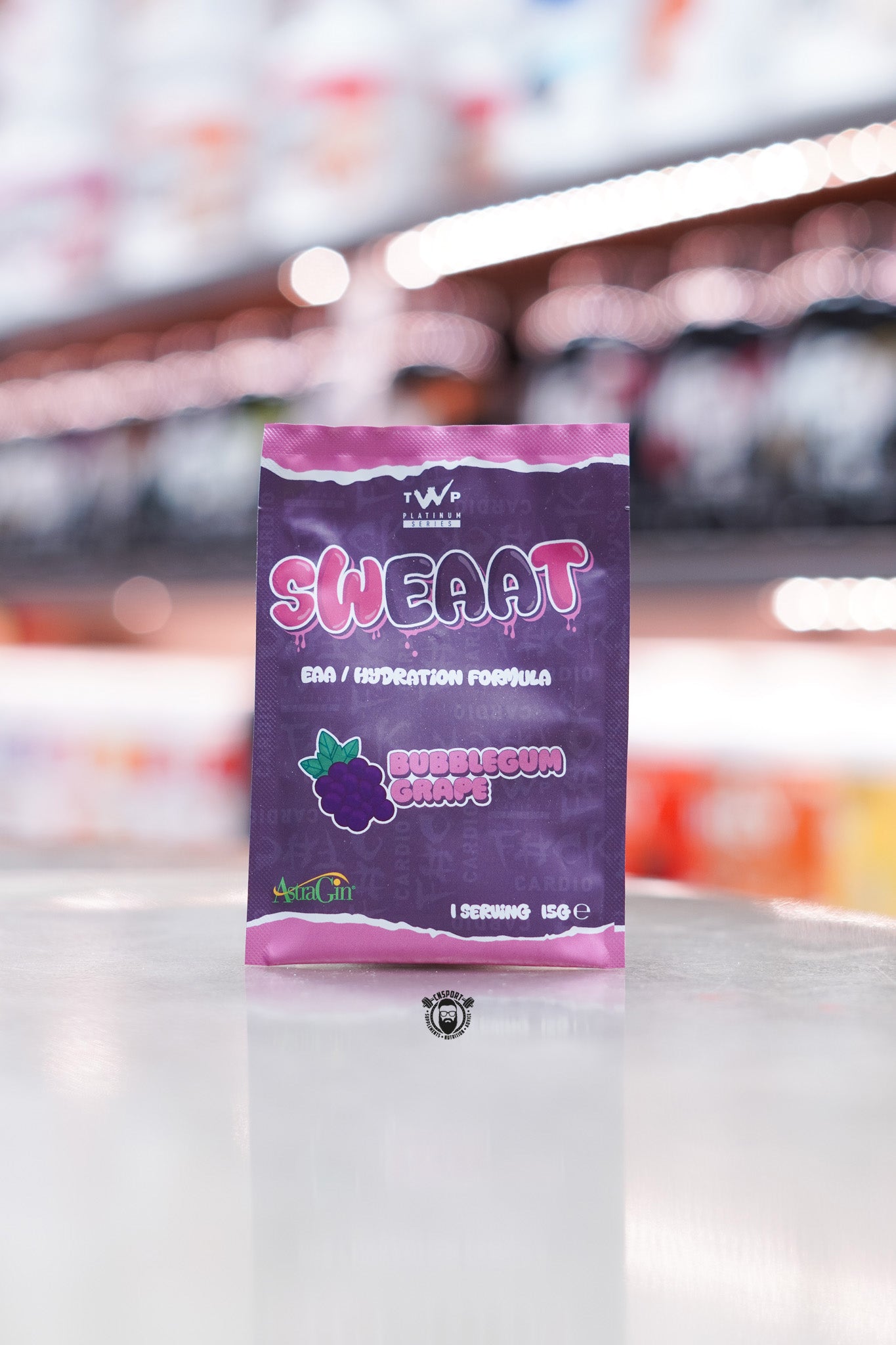 Single-serving 15g sample sachet in purple packaging with pink accents, featuring grape illustration and AstraGin logo, displayed upright on a store counter with supplement shelves blurred in the background.