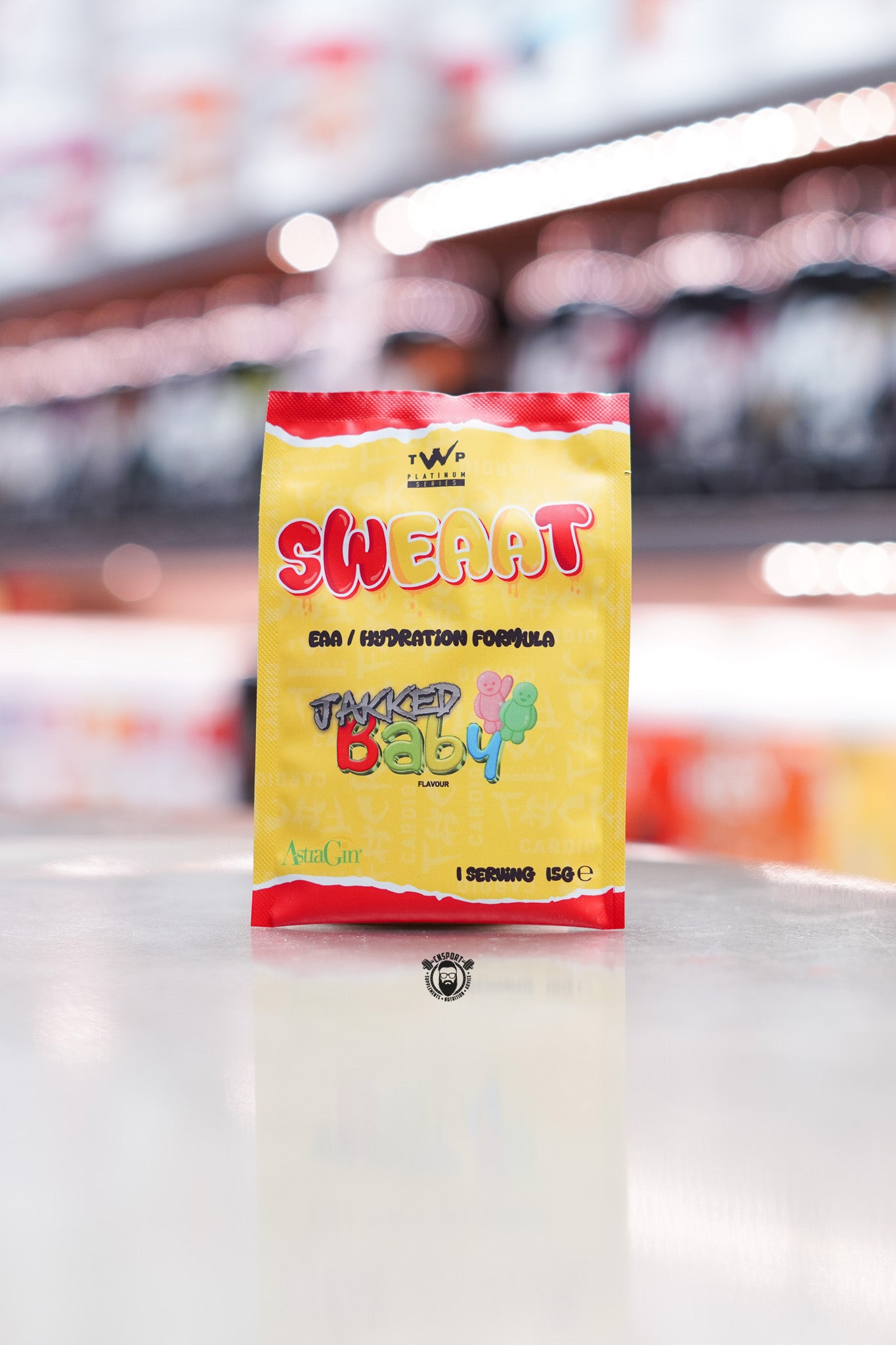Single-serving 15g sample sachet in yellow and red packaging with cartoon gummy bear illustration and AstraGin logo, standing on a store counter with blurred supplement shelving in the background.
